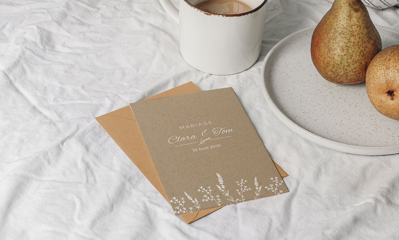 Photo d'un faire-part mariage posé sur une nappe blanche et sur une enveloppe kraft. Le faire-part est un grand format portrait au fond kraft et motifs végétaux blancs avec le nom des mariés et la date du mariage au centre.