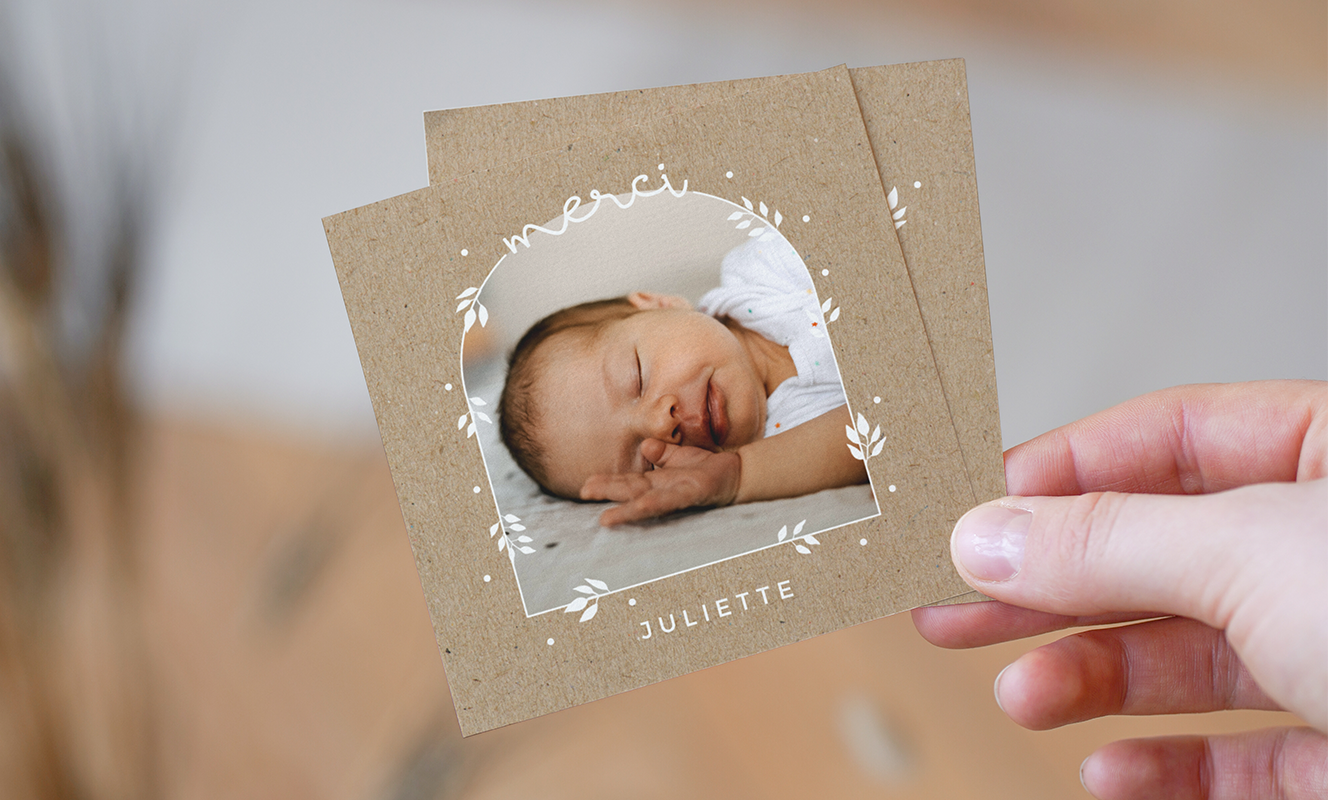 Photo d'une main qui tien une petite carte de remerciement naissance au format carré et fond kraft avec les motifs de végétaux blancs, une photo d'un bébé au centre au format arche, un texte merci sur le dessus et le prénom du bébé en dessous.