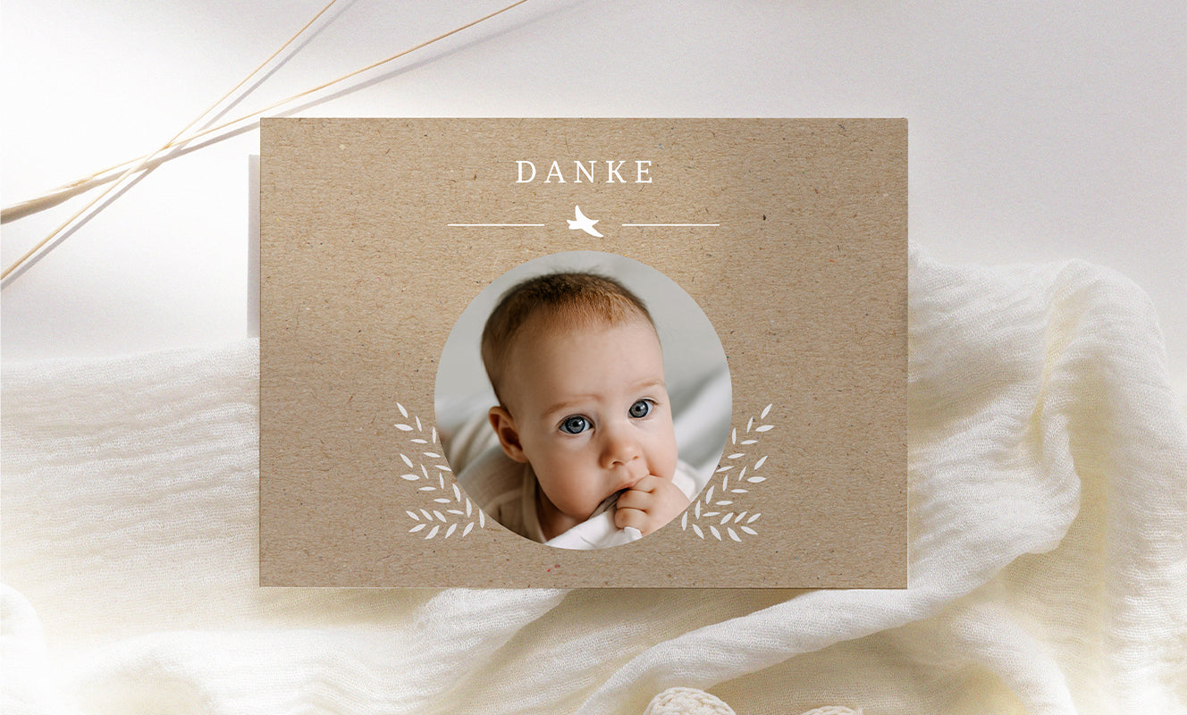 Foto einer einfachen Geburtsanzeige im Querformat, Krafthintergrund mit einem runden Babyfoto in der Mitte, einigen weißen Pflanzenmotiven und dem Wort „Danke“ über dem Foto.